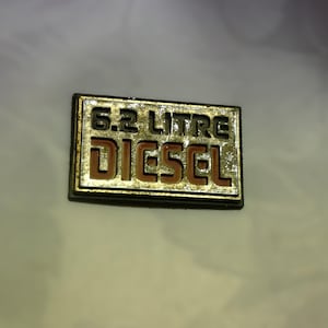 Puede incluir: Una placa de metal dorada con el texto "6.2 LITRE DIESEL" en letras negras y naranjas.