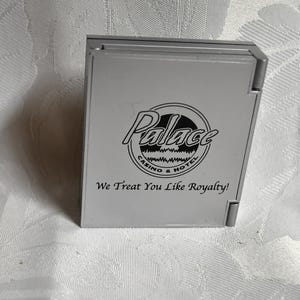 May include: A silver, hinged box with the word "Palace" in a circular logo, along with "Casino & Hotel". Below the logo, the text reads "We Treat You Like Royalty!"