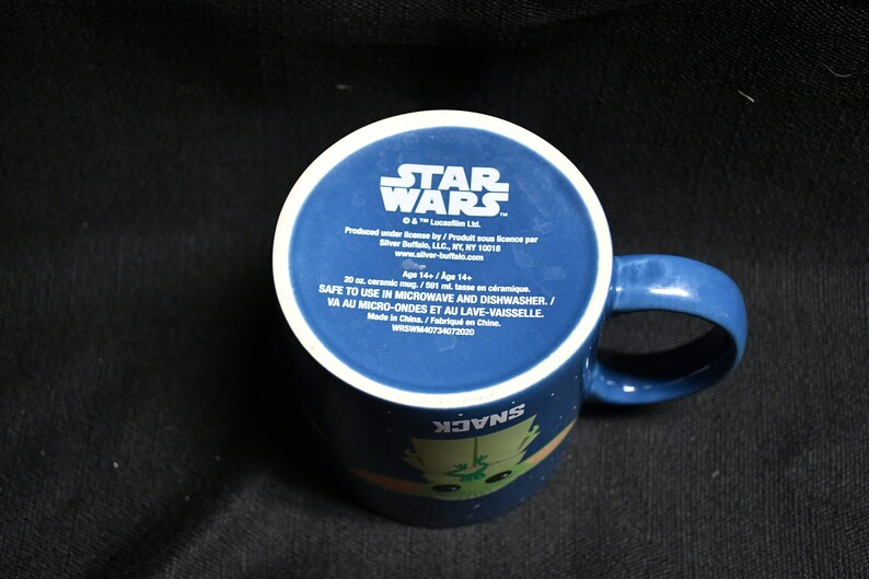 May include: A blue ceramic mug with a white rim and handle. The mug features a Star Wars logo and a cartoon image of a character from the movie. The text on the mug reads "Star Wars", "Safe to use in microwave and dishwasher", and "Made in China".
