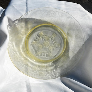 Può includere: Un piatto di vetro trasparente con bordo giallo e un design in rilievo dettagliato al centro. Il piatto è appoggiato su una tovaglia bianca, con la luce del sole che proietta ombre sulla superficie. Il design del piatto è floreale.
