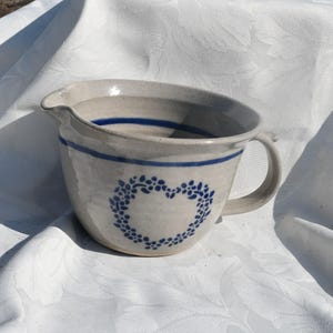 Puede incluir: Un cuenco de cerámica con asa y boquilla. El cuenco es de color gris claro con dos rayas azules y un diseño de corazón floral azul. El cuenco está sobre un mantel blanco.