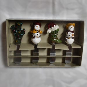 May include: Set of five Christmas themed cheese spreaders in a box. The spreaders feature a snowman, a cactus, a boot, and two cowboys.
