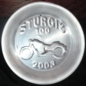 Könnte beinhalten: Ein Aschenbecher aus silbernem Metall mit den Worten "Sturgis 100 2003" und einer Motorrad-Silhouette.