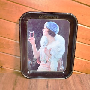 Puede incluir: Bandeja de servir Coca-Cola vintage con borde negro. La bandeja presenta a una mujer con turbante azul y chal de piel blanca sosteniendo un vaso de Coca-Cola. El logotipo de Coca-Cola está impreso en la parte superior de la bandeja.