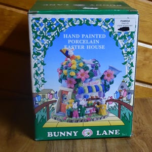 May include: A hand-painted porcelain Easter house shaped like a watering can with a bunny on top. The house has a small ice cream shop with a sign that says "Ice Shop". The house is surrounded by flowers and greenery. The box says "Bunny Lane" and "Hand Painted Porcelain Easter House".