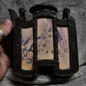 May include: A dark brown, antique lantern with three panels depicting a scene with figures. The lantern has a decorative top and base with ornate detailing. The panels have a faded, aged appearance, suggesting the lantern's age and history.