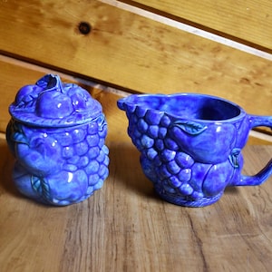 May include: A cobalt blue ceramic sugar bowl with a lid and a matching creamer. Both pieces are decorated with a grape cluster design. The sugar bowl is round, and the creamer has a handle.
