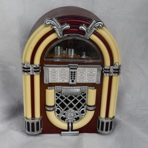 Könnte beinhalten: Eine Tisch-Jukebox mit einem Gehäuse in dunkler Holzoptik, cremefarbenen, bogenförmigen Verzierungen und silbernen Metallakzenten. Die Vorderseite zeigt ein Bedienfeld mit Songtiteln, ein zentrales Lautsprechergitter mit einem silbernen Emblem und ein transparentes Sichtfenster.