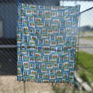 May include: A blue and white patterned fabric with a repeating design of cartoon characters, apples, and picnic scenes. The fabric is hanging on a clothesline, with a blurred background of a chain-link fence and greenery.