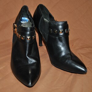 May include: A pair of black leather ankle boots with gold-toned studs on the straps. The boots have a pointed toe and a high heel. The size 6 1/2 is written on a blue tag.