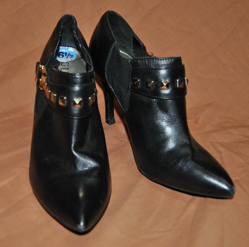 May include: A pair of black leather ankle boots with gold-toned studs on the straps. The boots have a pointed toe and a high heel. The size 6 1/2 is written on a blue tag.