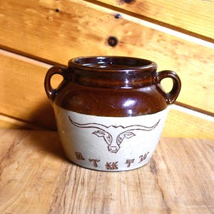 May include: A two-handled ceramic crock featuring a glossy dark brown glazed upper section and a matte light beige lower section. The front displays a brown line drawing of a longhorn steer head above a series of stylized brown cattle brand symbols.