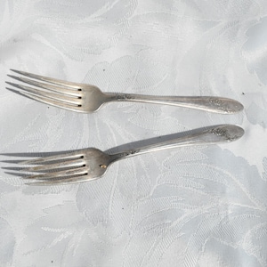 Könnte beinhalten: Zwei antike Silber-Gabeln mit kunstvollen floralen Details an den Griffen. Die Gabeln sind diagonal auf einer weißen, strukturierten Tischdecke angeordnet und zeigen ihre Zinken und das Gesamtdesign. Ein klassisches Set für das Essen.