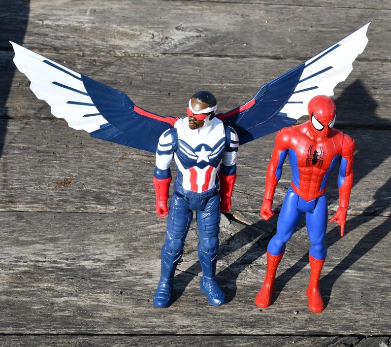 Puede incluir: Dos figuras de acci&oacute;n de superh&eacute;roes. Una figura viste un traje rojo, blanco y azul con una estrella y grandes alas blancas y azules. La otra figura lleva un traje rojo y azul con un emblema de ara&ntilde;a.