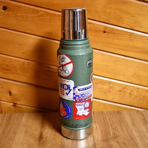 May include: A vintage green Stanley thermos with a silver lid and base. The thermos is covered in various stickers, including a "Union Yes" sticker and a "Buy American" sticker. The thermos is standing on a wooden surface.