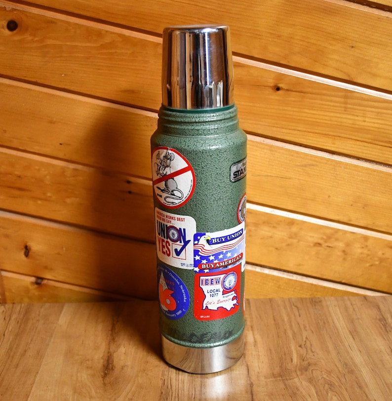May include: A vintage green Stanley thermos with a silver lid and base. The thermos is covered in various stickers, including a "Union Yes" sticker and a "Buy American" sticker. The thermos is standing on a wooden surface.