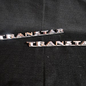 May include: Two silver-colored "TRANSTAR" emblems. The emblems are raised and have a slightly textured surface.