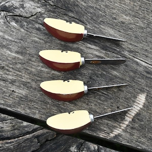 Peut inclure: Quatre couteaux à fromage en forme de pomme avec des lames en acier inoxydable. Les moitiés de pomme sont peintes avec un intérieur crème et un extérieur rouge, avec des détails de graines noires. Les couteaux sont disposés en diagonale sur une surface en bois vieilli.