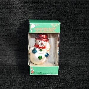 May include: A white glass snowman ornament with a red hat, blue mittens, and green accents. The ornament is in a clear plastic box with the text "COLLECTABLE BLOWN GLASS ORNAMENT" and "HO HO HO!!!".