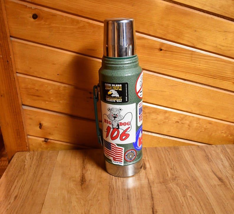 May include: A green and silver insulated thermos with a stainless steel lid. The thermos has several stickers, including one with a bulldog and the words "Big Dog 106" and another with an American flag.