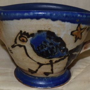 May include: A blue and white ceramic pitcher with a bird design. The pitcher has a handle and a small base. The bird is painted in black and blue with a yellow eye.
