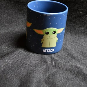 May include: A blue ceramic mug with a white star pattern and a green character with large ears. The character is wearing a brown robe and the text "ATTACK" is printed below it.