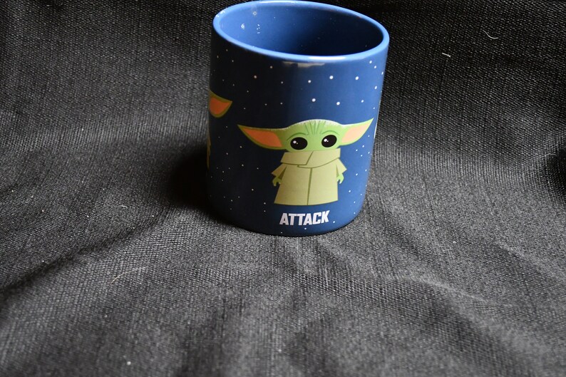 May include: A blue ceramic mug with a white star pattern and a green character with large ears. The character is wearing a brown robe and the text "ATTACK" is printed below it.
