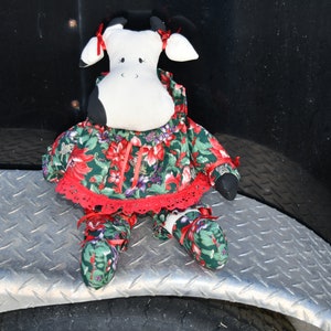 May include: A black and white cow plush toy wearing a red and green floral dress with a white lace trim. The toy is sitting on a silver metal surface.
