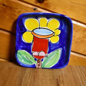 May include: A square ceramic plate with rounded corners featuring a hand-painted design of a stylized flower. The background is a deep blue, while the flower has yellow petals, a light blue center, a red stem, and two green leaves at the base.
