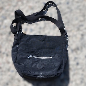 May include: Black crossbody bag with a long adjustable strap. The bag features a front zippered pocket with a silver zipper and a round logo patch. The bag is made of a durable, water-resistant material.