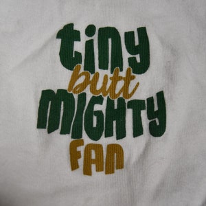 Op de afbeelding: Wit T-shirt met de tekst "tiny but mighty fan" in groene en gouden letters.