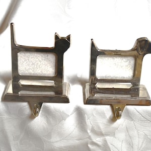 May include: Two silver-colored metal stocking holders, one shaped like a cat and the other a dog. Each has a rectangular frame for a photo and a hook for hanging. The cat has pointed ears, and the dog has a snout.