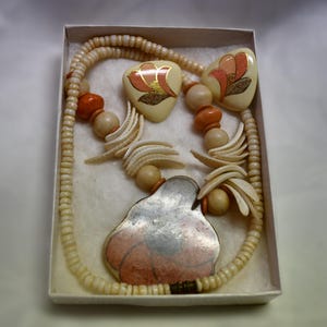 May include: A white and orange floral necklace and earrings set. The necklace is made of white beads, orange beads, and white shell-shaped beads. The earrings are heart-shaped with a white background and orange floral designs.