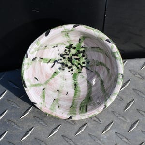 Könnte beinhalten: Eine handbemalte Keramikschale mit weißem Grund, die grüne und schwarze abstrakte Designs aufweist. Das Innere der Schale hat einen rosafarbenen Farbton, und der Rand ist mit schwarzen Pinselstrichen akzentuiert. Die Schale ist rund und scheint handgefertigt zu sein.