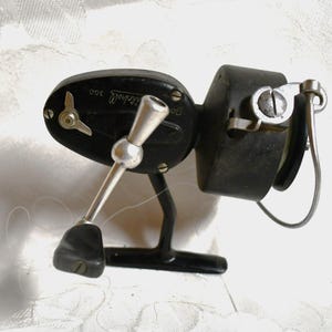 May include: A vintage Mitchell 300 fishing reel. The reel is primarily black with silver accents on the handle and other components. The reel is designed for use in fishing and has a classic, functional design.
