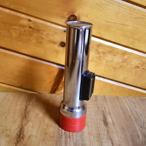 May include: A vintage chrome flashlight with a red base and a black switch. The cylindrical flashlight is made of shiny metal and sits on a wooden surface. The flashlight is a classic design.