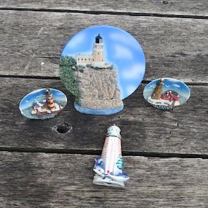 May include: A collection of lighthouse-themed decorative items. The items include a large circular piece with a lighthouse on a cliff, two oval pieces with lighthouse designs, and a small lighthouse figurine. The items are on a weathered wooden surface.