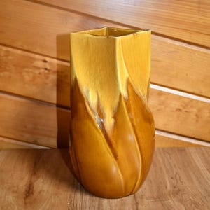 May include: A brown and yellow ceramic vase with a unique, wavy design. The vase has a square opening at the top and a rounded bottom.