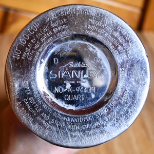 May include: Close-up of the bottom of a vintage Stanley vacuum bottle. The silver-colored metal base features the Aladdin and Stanley logos, along with text indicating the model number, "NO A-944DH QUART", and instructions for use.