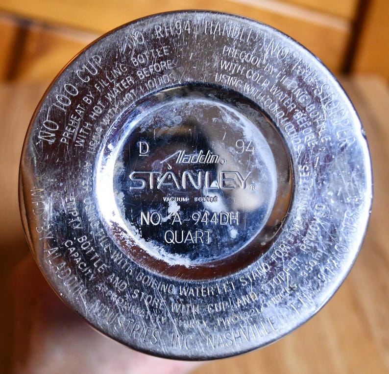 May include: Close-up of the bottom of a vintage Stanley vacuum bottle. The silver-colored metal base features the Aladdin and Stanley logos, along with text indicating the model number, "NO A-944DH QUART", and instructions for use.