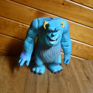 May include: A blue monster action figure with purple spots, two curved yellow horns, and a wide grin showing white teeth. The figure has a textured, fur-like appearance, articulated arms, and a speaker grille located on the stomach area.