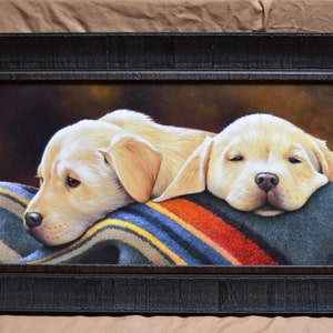 May include: Two yellow Labrador Retriever puppies are napping on a colorful striped blanket. The puppies are lying side by side with their heads resting on the blanket. The painting is framed in a dark wood frame.
