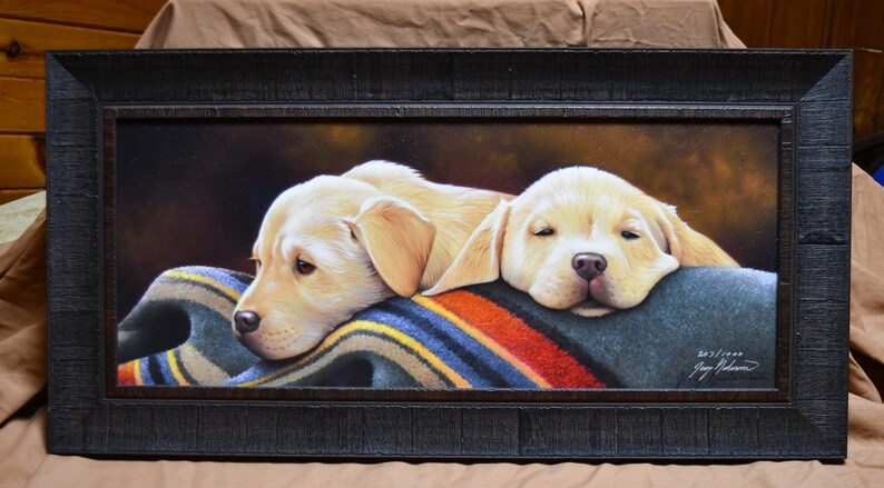 May include: Two yellow Labrador Retriever puppies are napping on a colorful striped blanket. The puppies are lying side by side with their heads resting on the blanket. The painting is framed in a dark wood frame.