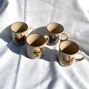 May include: Four beige ceramic mugs with brown rims and handles. Each mug features a different butterfly design with colorful wings and floral accents. The mugs are arranged on a white surface.