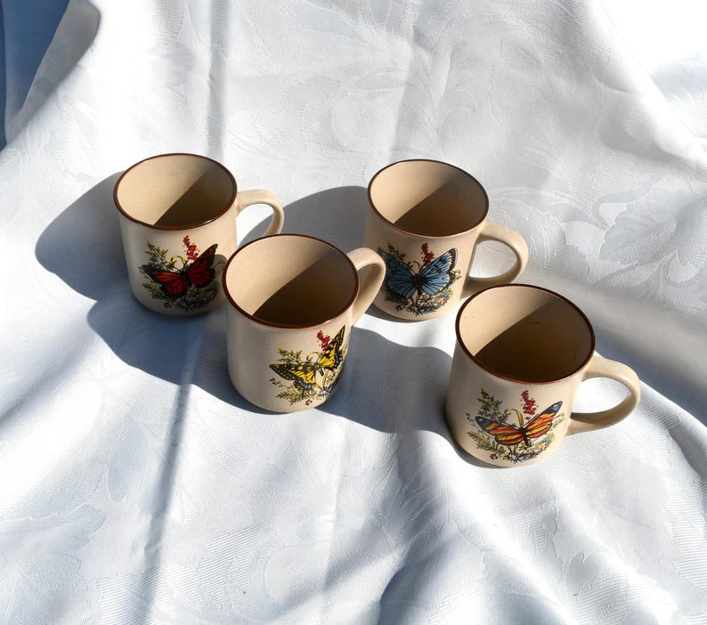 May include: Four beige ceramic mugs with brown rims and handles. Each mug features a different butterfly design with colorful wings and floral accents. The mugs are arranged on a white surface.