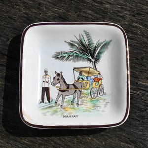 May include: Square ceramic dish with a detailed illustration of a horse-drawn carriage, palm tree, and a person in uniform. The word "NASSAU" is printed at the bottom. The dish has a white background with a brown border.