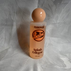 May include: A vintage Faberge Babe Splash Cologne bottle. The clear glass bottle contains a peach-coloured liquid. The bottle has a peach-coloured ball-shaped cap. The label features the word "Babe" in an orange circle and the words "Splash Cologne".