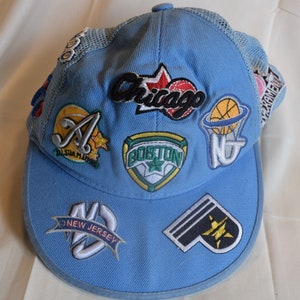 May include: A light blue baseball cap with a mesh back and various embroidered patches. The patches include logos for the Atlanta Braves, Boston Red Sox, Chicago Bulls, New Jersey Nets, and a star.