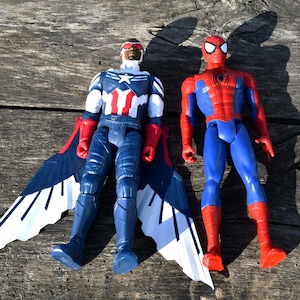 Puede incluir: Dos figuras de acci&oacute;n de superh&eacute;roes. La figura de Falcon viste un traje azul, blanco y rojo con grandes alas. La figura de Spider-Man viste un traje rojo y azul. Ambas figuras est&aacute;n sobre una superficie de madera desgastada.