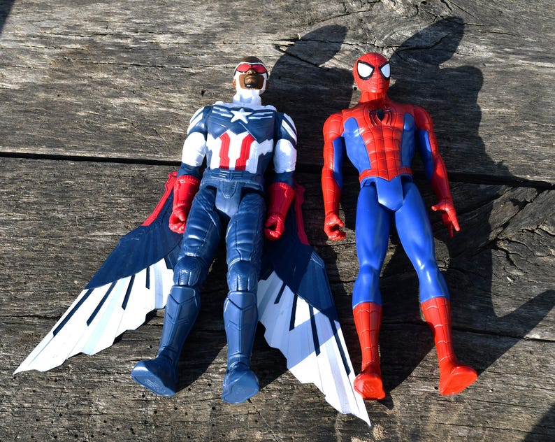 Puede incluir: Dos figuras de acci&oacute;n de superh&eacute;roes. La figura de Falcon viste un traje azul, blanco y rojo con grandes alas. La figura de Spider-Man viste un traje rojo y azul. Ambas figuras est&aacute;n sobre una superficie de madera desgastada.